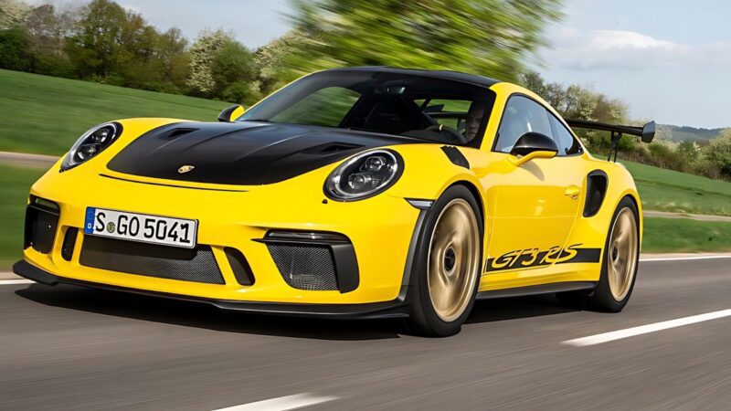 A yellow Porsche 911 GT3 RS with striking black accents speeds along a scenic road, surrounded by lush greenery and trees in the background.