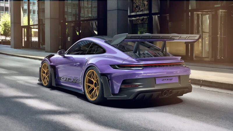 A striking Porsche 911 GT3 RS in purple, featuring gold wheels and a prominent rear wing, is parked on a city street surrounded by sleek glass buildings.