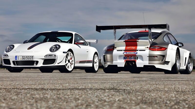 Two Porsche 911 GT3 RS cars, one facing forward and the other shown from the rear, are parked on an open pavement under a partly cloudy sky, showcasing the striking design and performance of the Porsche 911 GT3 RS.