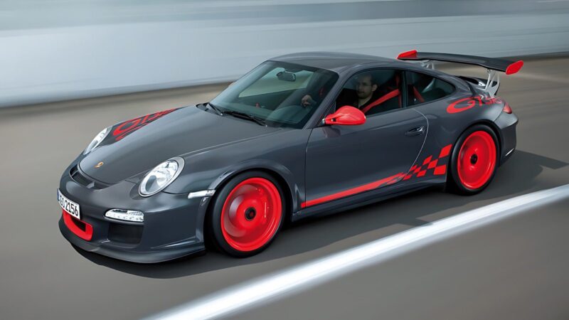 A gray Porsche 911 GT3 RS with striking red wheels and accents is driving on a road, captured from above and the side.