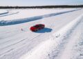 A sleek Ferrari 296 GTB zips along a snowy track, with a single orange cone marking the way, surrounded by a pristine snow-covered landscape under a clear blue sky.
