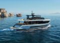 A modern white Arcadia Yachts A80new cruises on calm blue water near rocky outcrops under a clear sky, with distant mountains in the background, blending luxury with a commitment to sustainability.
