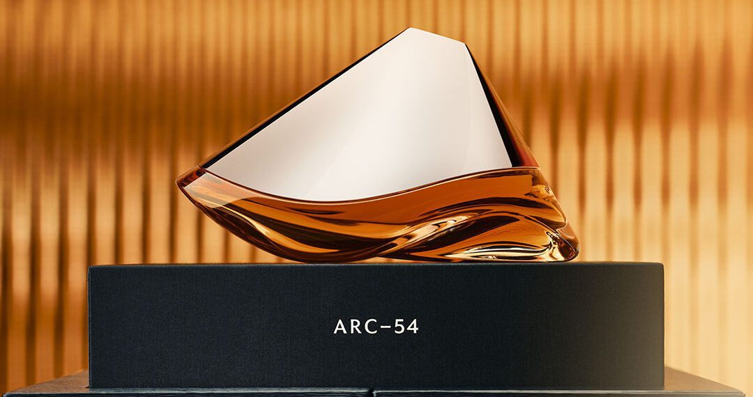 A sleek, geometrically shaped glass bottle of Arc-54 perfume sits on a black box against a gold, textured background, reminiscent of the elegance and precision of an Aston Martin Valkyrie Hypercar.