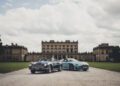 Two vintage Aston Martins parked on a gravel path in front of an elegant, large mansion under a cloudy sky, embodying the timeless grace befitting a setting that whispers of a New Royal Warrant.