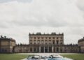 Two luxury cars, including an Aston Martin, are elegantly parked on a gravel driveway in front of a large historic building under a partly cloudy sky, exuding an air of prestige befitting a Royal Warrant.