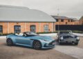 A modern blue convertible Aston Martin is parked beside a classic black car, embodying the elegance deserving of a Royal Warrant, in front of a brick building.