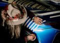 A person with long hair and a hat decorated with flowers applies masking tape to the rear quarter panel of a blue Magnus Walker car with white stripes.