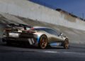 A metallic sports car with a large rear spoiler and bronze wheels, inspired by a collaboration with Magnus Walker, is parked on a paved surface next to a concrete embankment.