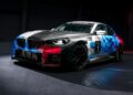 A BMW M2 Racing car with a digital-patterned, multicolored livery is parked indoors under a spotlight, proudly displaying racing number 1 on its door.