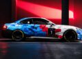 A BMW M2 Racing car with a geometric blue, red, and white livery is parked indoors under dramatic lighting, the number 1 boldly displayed on the door.
