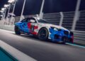 A BMW M2 Racing car with a blue, red, and white geometric pattern drives on a brightly lit track at night.