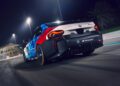 A BMW M2 Racing car with a blue, red, and white livery speeds around the racetrack at night under bright lights.