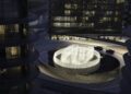 A modern roundabout in Abu Dhabi features a large circular fountain illuminated at night, surrounded by glass buildings and two cars driving nearby—a scene evocative of the city's luxury real estate.