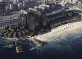 Aerial view of BRABUS Island, a luxury real estate beachfront resort in Abu Dhabi with high-rise buildings, villas, sandy beach, umbrellas, and a marina with yachts along a curved shoreline.