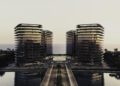 Two identical modern glass buildings, part of BRABUS Island luxury real estate in Abu Dhabi, are connected by a central structure over a road, with water on both sides and the sea glowing at sunset in the background.