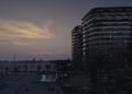 Modern beachfront high-rise buildings at dusk in Abu Dhabi, with lights shining from luxury real estate overlooking a calm bay and a sandy beach dotted with lounge chairs and palm trees.