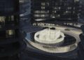A circular, illuminated fountain is surrounded by modern buildings and luxury real estate, with cars parked along a curved driveway at night on BRABUS Island, Abu Dhabi.