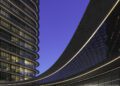 Modern, curved glass building with illuminated lines at dusk on BRABUS Island in Abu Dhabi; people are visible near the entrance and a black vehicle is parked in front, showcasing true luxury real estate.