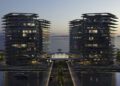 Two modern glass high-rise buildings with illuminated windows stand by the water at dusk in Abu Dhabi, flanking a circular entrance with a central fountain and a bridge leading to this luxury real estate complex.