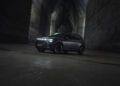 A dark-colored SUV parked in a dimly lit, cave-like setting with rugged stone walls and a rough floor. The vehicle's headlights are on, illuminating the surroundings.