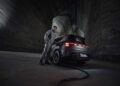 A man in a jumpsuit stands beside a sleek, gray electric car boasting 637HP, charging in a dimly lit, cavernous space with textured walls.