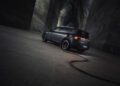 A dark SUV, possibly a BRABUS 637HP, is parked in a dimly lit industrial space with a cable plugged into it, indicating charging. The vehicle's rear lights glow softly, casting an intriguing light on the textured walls around.