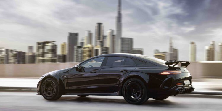 A BRABUS Rocket 1000, a luxury car, speeds along a city road with towering skyscrapers and a tall tower in the background beneath a cloudy sky.