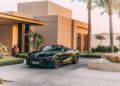 A black luxury convertible car is parked in the driveway of a modern house with palm trees and landscaped gardens in the background.