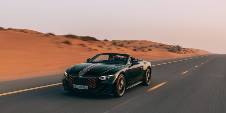 A dark green Bentley Batur Convertible makes its public debut, driving on an empty road through a desert landscape at sunset near Dubai.