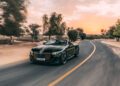 The Bentley Batur Convertible, a green luxury sports car with orange accents, makes its public debut as it drives on a curving road at sunset, surrounded by trees and Dubai’s desert landscape.