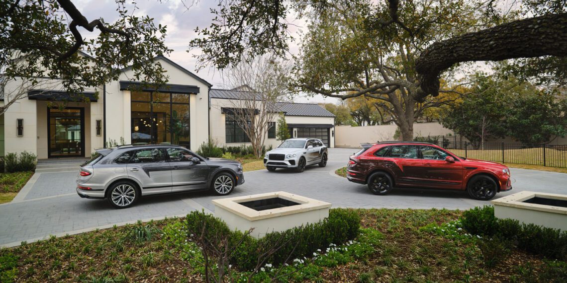 Three luxury SUVs, including bespoke Victoris Bentaygas in silver, white, and red, are parked on a paved driveway. They stand in front of a modern house with large windows, surrounded by trees and lush landscaping.