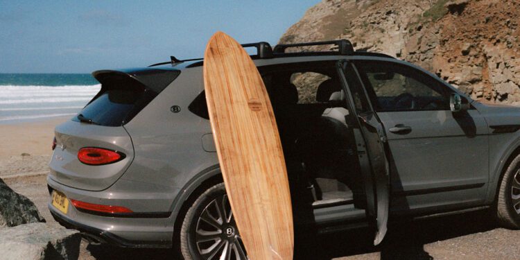 A wooden Otter Surfboards surfboard is propped against a grey SUV parked near a rocky beach with open car doors; the ocean and cliffs are visible in the background, hinting at an inspiring surfboard collaboration.