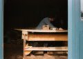 A person works barefoot at a wooden table inside the Otter Surfboards workshop, using tools and materials, seen through an open blue door—a glimpse into a unique surfboard collaboration.