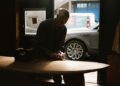 In a dimly lit workshop, a craftsman sands an Otter Surfboards creation, hinting at a unique surfboard collaboration. A Bentley and an open door are visible in the background, adding to the scene’s refined atmosphere.