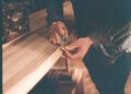 A person uses a hand planer to smooth the edge of a wooden board on a workbench, much like the careful craftsmanship seen in Otter Surfboards' surfboard collaboration projects.