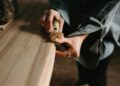 A person uses a hand tool to shave and smooth the edge of a wooden surface, with wood shavings curling from the blade—crafting what could be an Otter Surfboards creation or part of a unique surfboard collaboration.