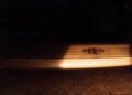 A wooden surface with a visible oval-shaped mark, reminiscent of an Otter Surfboards and Bentley surfboard collaboration, is partially illuminated by a beam of light, while the background remains mostly in shadow.