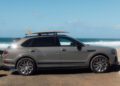 A gray Bentley SUV with a surfboard on the roof rack, crafted through an Otter Surfboards collaboration, is parked near a beach with waves and blue sky in the background.