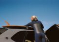 A person in a wetsuit places an Otter Surfboards surfboard on the roof rack of a parked Bentley under a clear blue sky.