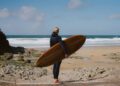 Person in a wetsuit holding an Otter Surfboards wooden surfboard stands on a rocky beach, facing the ocean under a partly cloudy sky.