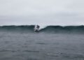A person is surfing on a wave under an overcast sky atop an Otter Surfboards x Bentley surfboard, with the ocean appearing calm in the foreground.