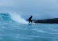 A person in a wetsuit is surfing a wave near the shore under cloudy skies, riding an Otter Surfboards and Bentley surfboard collaboration.