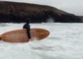A person in a wetsuit stands in the ocean holding a Bentley-collaboration wooden surfboard, looking toward rocky cliffs under an overcast sky.