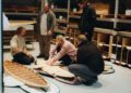 Five people examine wooden surfboard pieces on the floor in a workshop with shelves holding wooden materials—part of an Otter Surfboards and Bentley surfboard collaboration.
