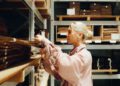 A person with blonde hair and a pink shirt organizes flat wooden sheets, part of an Otter Surfboards collaboration, on labeled shelves in a storage area.