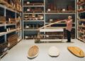 A person examines a wooden plank in a workshop, surrounded by stacks of wood boards and finished items, including an Otter Surfboards x Bentley surfboard collaboration displayed on the floor.
