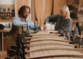 Two people in a woodworking shop use clamps and apply glue to long wooden planks, collaborating on an Otter Surfboards project surrounded by tools and materials.
