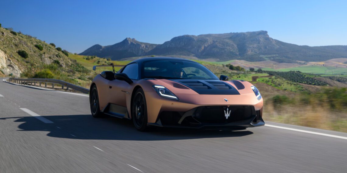 A sleek Maserati sports car races along a highway with mountainous scenery in the background under a clear blue sky.