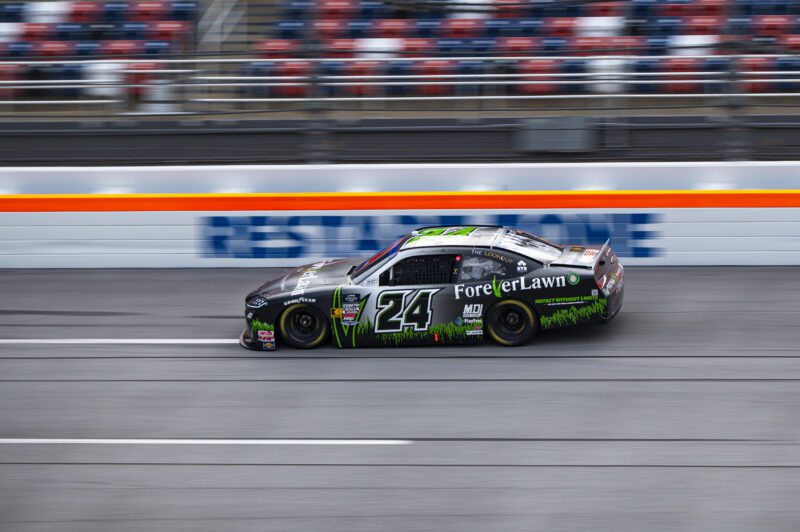 A black and green standard race car with the number 24 and "ForeverLawn" branding speeds down a racetrack, aiming for the top, in front of empty stadium seats.