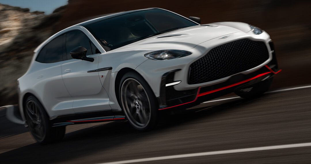 A white Aston Martin DBX S Super-SUV with black and red accents is driving at speed on a curved mountain road.